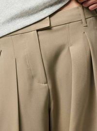 Beežid pehmes kangas lõhikutega püksid, millel on küljetaskud ja kohandatud vöökoht, esitletud halli ülepingutusega.