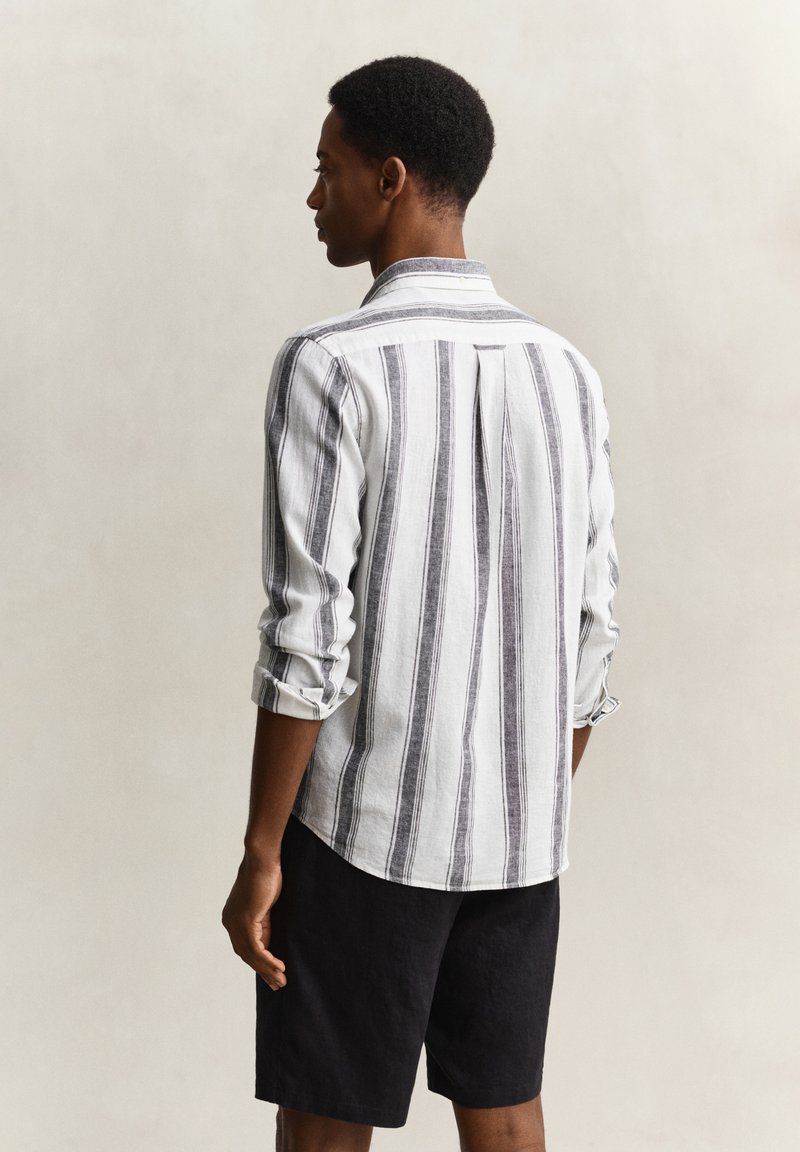 Man met kort haar die een wit shirt met lange mouwen en zwarte verticale strepen en zwarte shorts draagt, met de rug naar de kijker toe, tegen een effen achtergrond.