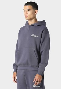 Šedá mikina z měkké tkaniny, se přední kapsou, žebrovanými manžetami a logem na hrudi. Spárováno s odpovídajícími šedými tepláky.