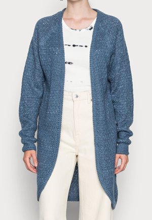 Personne portant un long cardigan bleu en maille, un haut blanc tie-dye et un pantalon crème taille haute, debout devant un fond uni.
