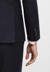 Miehen käsi siniraitaiseen pukutakin hihaan vieressä, jossa on neljä nappia, ja näkyvissä vaaleanpunainen paidan kalvosin.