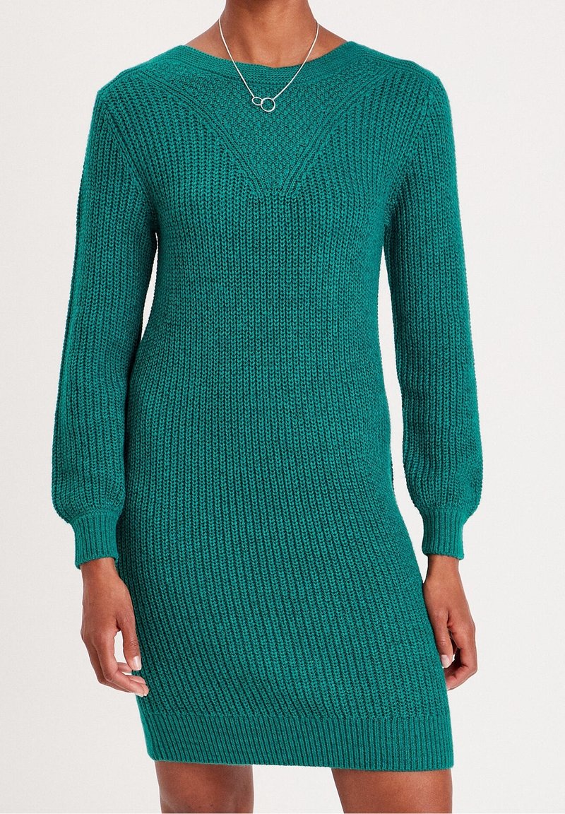 Femme portant une robe-pull tricotée bleu sarcelle à manches longues avec un col rond et un collier argenté avec deux cercles reliés.