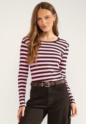 Donna con capelli lunghi castano chiaro indossa una maglia a maniche lunghe a righe orizzontali rosa e marrone scuro, pantaloni neri e una cintura marrone scuro con fibbia.