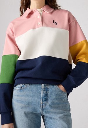 Henkilö, joka pukeutunut värilohkorugbypaitaan, jossa on vaaleanpunaisia, kermaisia, laivastonsinisiä, vihreitä ja keltaisia osioita, ja vaaleansinisiin farkkuihin, kädet taskuissa.