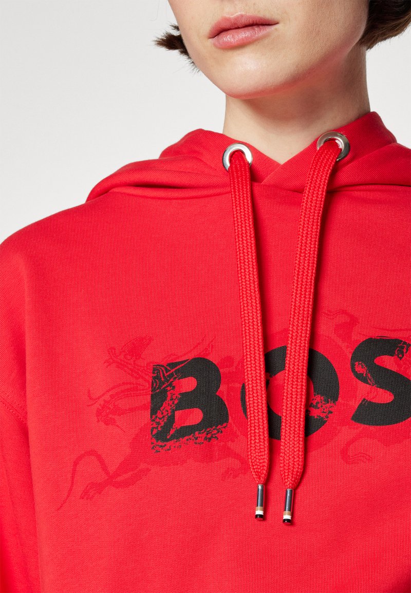 Vue partielle d'une personne portant un hoodie rouge vif avec des cordons épais et le texte "BOSS" noir en dégradé sur la poitrine.