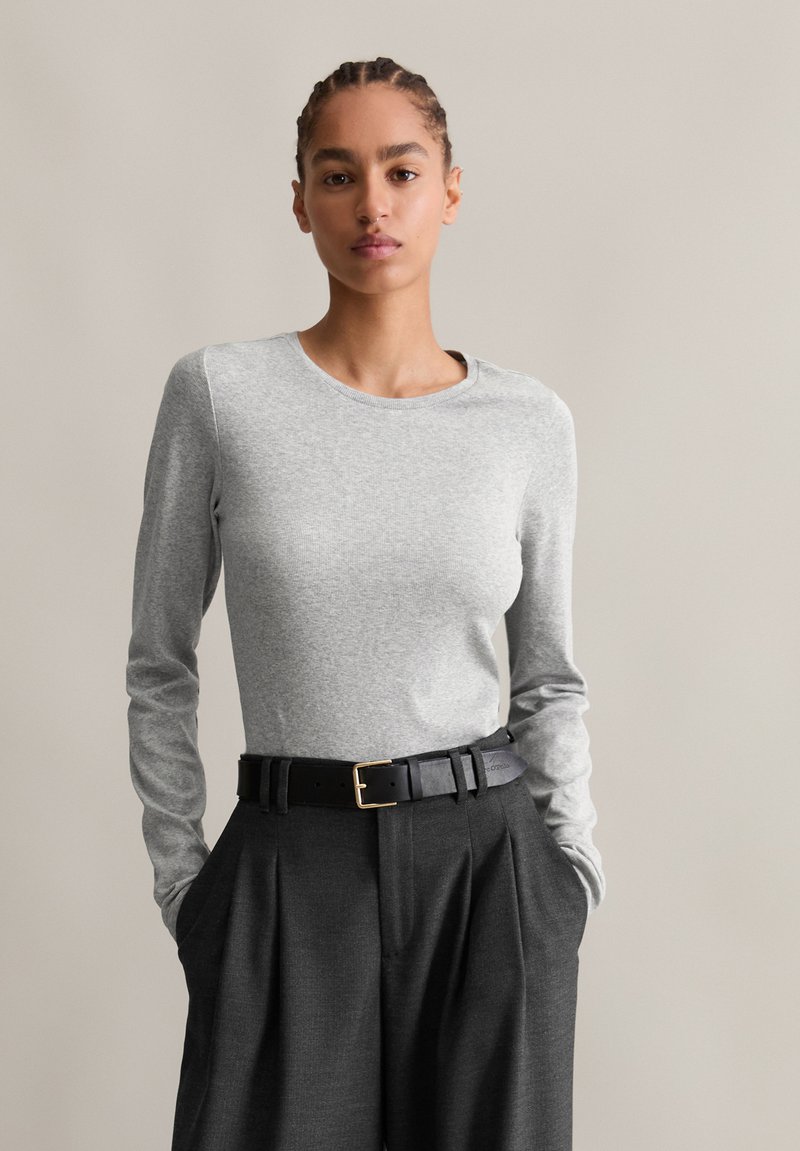 Világosszürke, hosszú ujjú póló kerek dekoltárral, magas derekú sötét nadrággal és fekete bőr övvel, arany csattal párosítva.