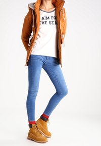 Brun huva jacka med dragsko över en vit skjorta med svart text, i kombination med blå tajta jeans och beige ankelstövlar. Röda strumpor synliga.