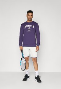 Lila pulóver "ADIDAS TENNIS CLUB" felirattal, fehér rövidnadrág lila díszítéssel, fekete cipő, teniszütőt tartva. Egyszerű háttér.