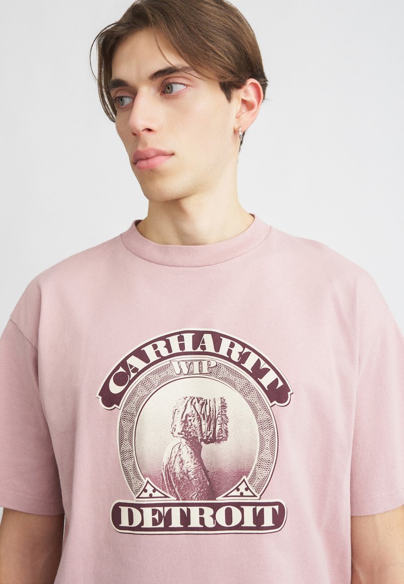 Roza bombažna majica s kratkimi rokavi, ki vsebuje grafični tisk profila figure z napisom "CARHARTT WIP DETROIT" v temno maroon barvi. Klasičen kroj.