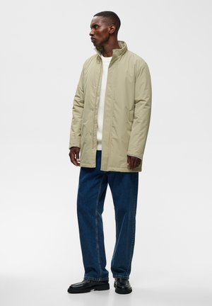 Homme portant une veste beige, un pull blanc, un jean bleu et des chaussures noires, debout devant un fond blanc, regardant sur le côté.