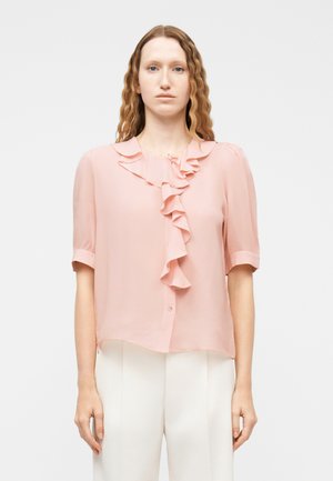 Žena s vlnitými červenými vlasmi má na sebe svetloružovú blúzku so záhybmi a biele nohavice, stojí na jednoliatom svetlom pozadí.