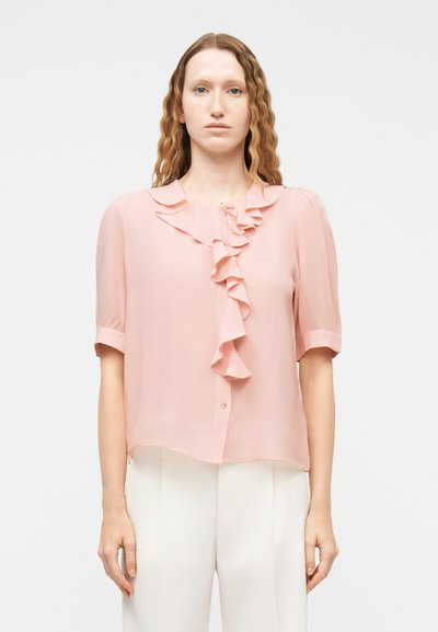 Femme aux cheveux roux ondulés portant un chemisier rose clair à volants et un pantalon blanc, debout devant un fond clair uni.