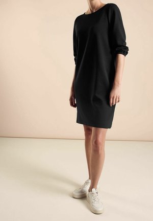 Kobieta w czarnej sukience z długim rękawem do kolan, nosząca białe trampki, na tle jednolitego beżowego tła.