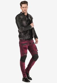 Schwarze Lederjacke mit gerippter Textur und Nieten, kombiniert mit strukturierten burgunderroten Jeans, die Reißverschlüsse und schwarze Lederakzente aufweisen.