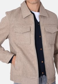 Giacca in misto lana beige con zip frontale, due tasche sul petto e colletto. Indossata sopra una camicia blu a bottoni.