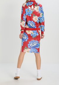 Röd blommig tracksuit-jacka med blå blommor, matchande kjol, tre vita ränder på sidorna, tillverkad av mjukt tyg, sportig design.