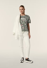 Camicia a maniche corte verde e bianca a fantasia, pantaloni della tuta bianchi e scarpe da ginnastica nere. Il modello tiene in mano una giacca bianca e si trova su uno sfondo neutro.