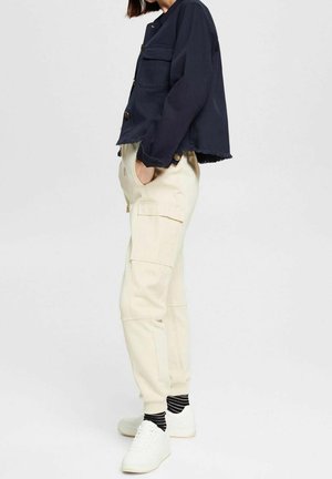 Persoon draagt een marineblauw cropped jasje, crèmekleurige cargobroek met zakken, zwart-wit gestreepte sokken en witte sneakers, staand met handen in de zakken.