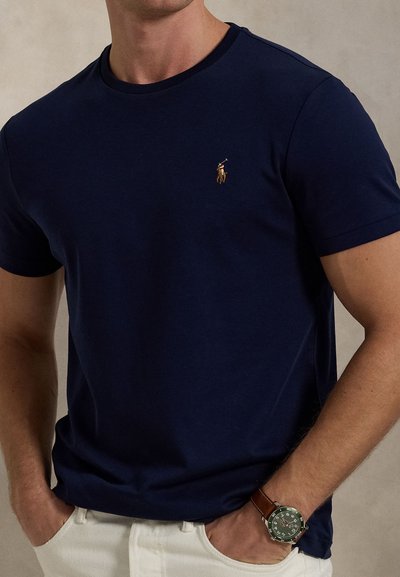 Moški nosi temno modro majico s logotipom igralca polo na prsih, bele hlače in rjave usnjene ure s zelenim številčnico, roke v žepih.