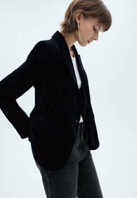 Sort fløjl blazer med spidsede klapper, polstrede skuldre og sidelommer, parret med en hvid skjorte og mørke jeans. Teksturstoffet er synligt.