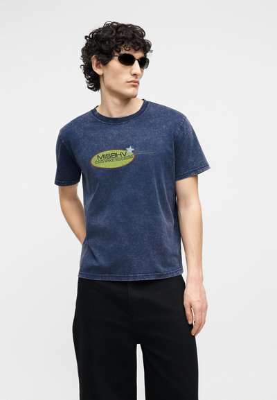 Άνδρας με σγουρά μαλλιά που φοράει μαύρα γυαλιά ηλίου, μπλε σκούρο φανερωμένο μπλουζάκι με στάμπα και μαύρο παντελόνι, στέκεται με το ένα χέρι πίσω από την πλάτη.