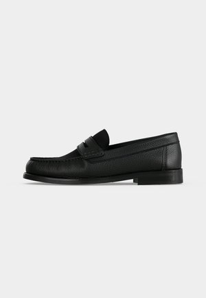 Mocassin penny en cuir noir avec surface texturée, petit talon et une lanière sur le dessus, présenté de profil sur un fond uni.