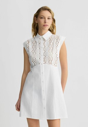 Abito bianco con scollo a polo e maniche a raglan. Presenta dettagli in pizzo intricati nella parte superiore e chiusura con bottoni lungo il davanti.