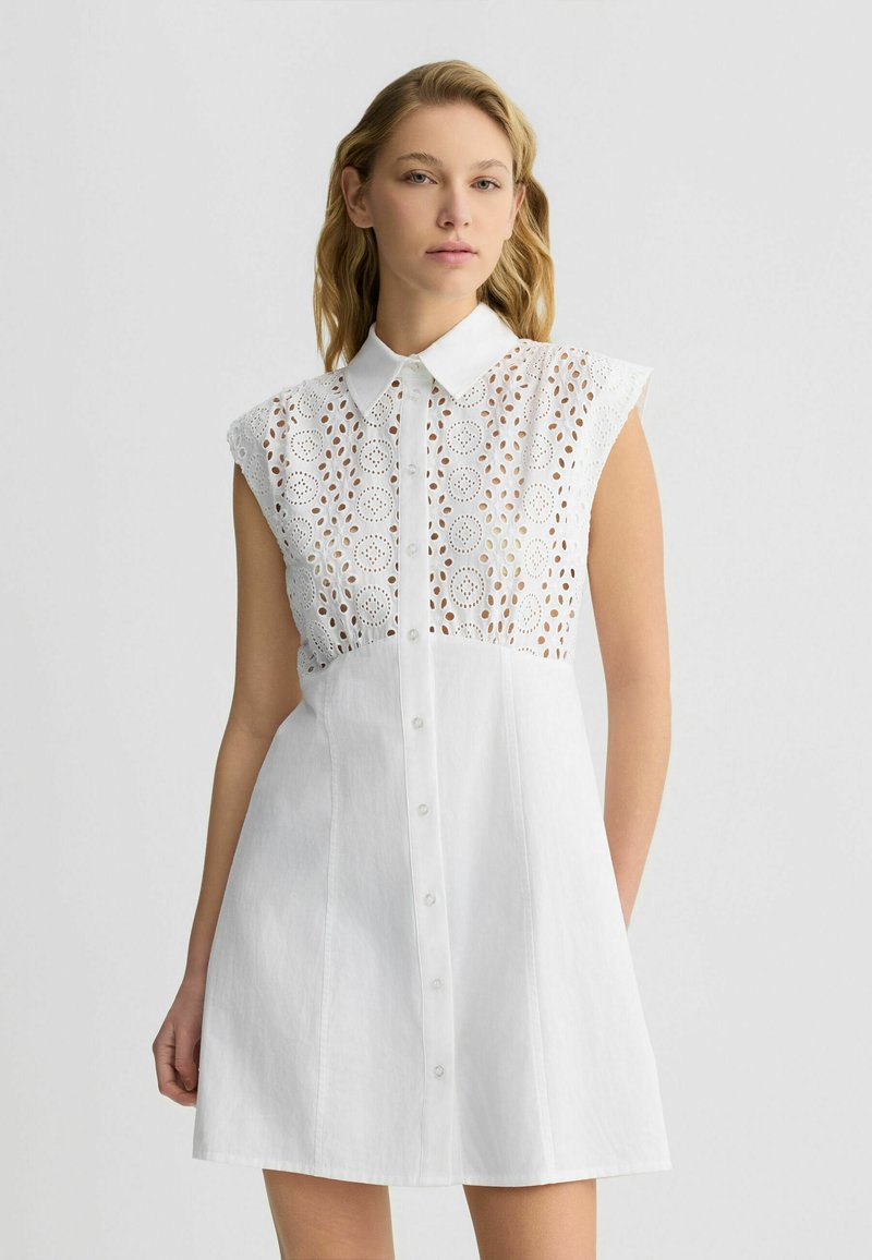 Abito bianco con scollo a polo e maniche a raglan. Presenta dettagli in pizzo intricati nella parte superiore e chiusura con bottoni lungo il davanti.