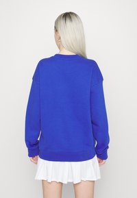 Blå sweatshirt i mjukt material, med rund halsringning, sänkta axlar och ribbade muddar. Bärs över en vit plisserad kjol.