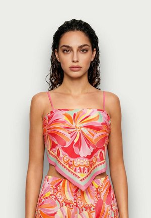 Femme aux cheveux bouclés mouillés portant un haut foulard rose à motif floral et un pantalon assorti, debout devant un fond uni clair.