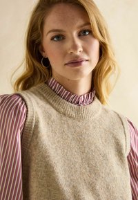 Beige gestrickte Pulloverweste mit einem gerippten Ausschnitt über einer pink-weiß gestreiften Bluse mit Rüschenkragen und Puffärmel.
