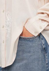 Blusa leve rosa, transparente, com bordado branco na frente, punhos com botões e botões semelhantes a pérolas, combinada com jeans azul.