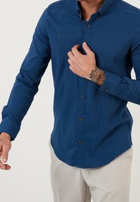 Niebieska koszula z długim rękawem w wzory, z kołnierzykiem zapinanym na guziki, teksturowana tkanina i ciemne guziki, noszona na jasnych spodniach.