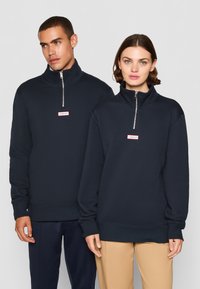 Felpe mezza zip blu navy realizzate in tessuto misto cotone; con polsini a costine e una piccola toppa con logo in rosso e bianco sul petto.