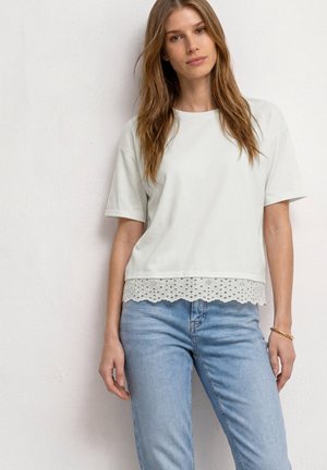 Femme aux longs cheveux portant un haut blanc à manches courtes avec bordure en dentelle et un jean bleu clair, debout contre un mur blanc.