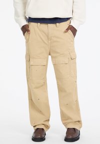 Beżowe spodnie cargo z wieloma kieszeniami, luźny krój i widocznymi plamami farby. Stylizowane z brązowymi butami.