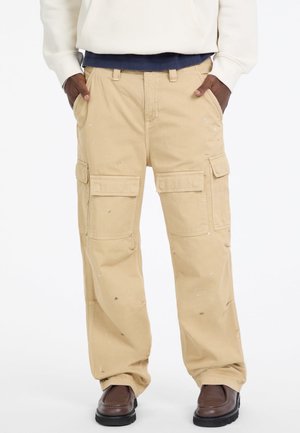 Beżowe spodnie cargo z wieloma kieszeniami, luźny krój i widocznymi plamami farby. Stylizowane z brązowymi butami.