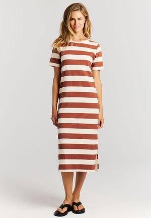 Vrouw staand, gekleed in een midi-jurk met horizontale strepen in bruin en wit, korte mouwen, en zwarte sandalen met open tenen, tegen een effen achtergrond.