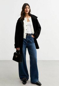 Cappotto oversize nero, cardigan bianco con bottoni davanti e jeans blu a gamba larga. Completano il look una borsa nera e mocassini.