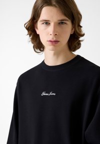 Czarna teksturowana bluza z okrągłym dekoltem, z białym haftem "Guess Jeans" na piersi. Luźny krój i długie rękawy.