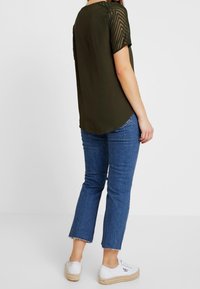 Olivenfarvet top med korte, teksturerede mesh-ærmer, parret med beskårne blå jeans og hvide sneakers. Enkel design og afslappet pasform.