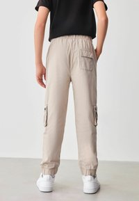 Beige Cargo-Hosen mit elastischem Bund, Seitentaschen und eng anliegenden Bündchen. Der Stoff hat eine glatte Textur und wird mit weißen Sneakers kombiniert.