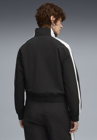 Czarna kurtka z białymi paskami po bokach, z wysokim kołnierzem, elastycznym paskiem w talii i teksturowanym materiałem. Widok z tyłu.