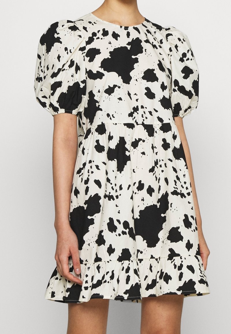 Robe courte imprimé vache noir et blanc avec manches bouffantes et ourlet à volants portée par une personne avec un bras détendu le long du corps.
