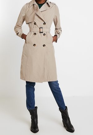 Femme portant un trench-coat beige croisé avec ceinture, un jean bleu et des bottines noires, debout les mains dans les poches du manteau.