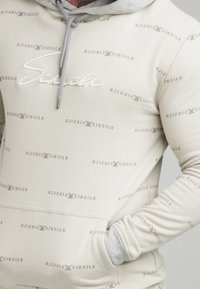 Sudadera color beige con detalles en gris, presenta un patrón de logo repetido, cierre con cordón y un bolsillo frontal. Fabricada en tela suave.
