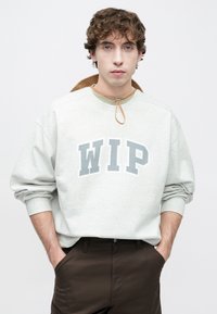 Γκρι φούτερ με στάμπα «WIP» σε σκούρο γκρι, ελαστικές μανσέτες και ελαστικό λαιμόκοψη. Φορεμένο πάνω από σκούρο καφέ παντελόνι. Το μοντέλο έχει ατημέλητα μαλλιά.