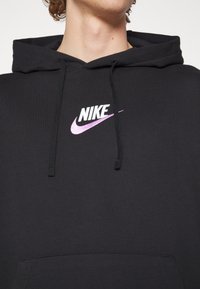 Svart hoodie med en ficka fram. Har en vit och lila Nike-logga centrerad på bröstet. Dragsnören i matchande svart tyg.