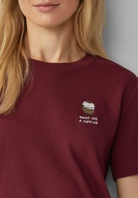 Bordeaux katoenen t-shirt met een cupcake borduursel en de tekst "SWEET LIKE A CUPCAKE" in het wit, met een ronde hals en korte mouwen.