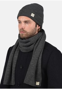 Pelēka adīta cepure un saskaņota šalle, abas ar ribotu faktūru un birku. Šalle brīvi krīt ap kaklu un ir kombinēta ar tumšu mēteļu.